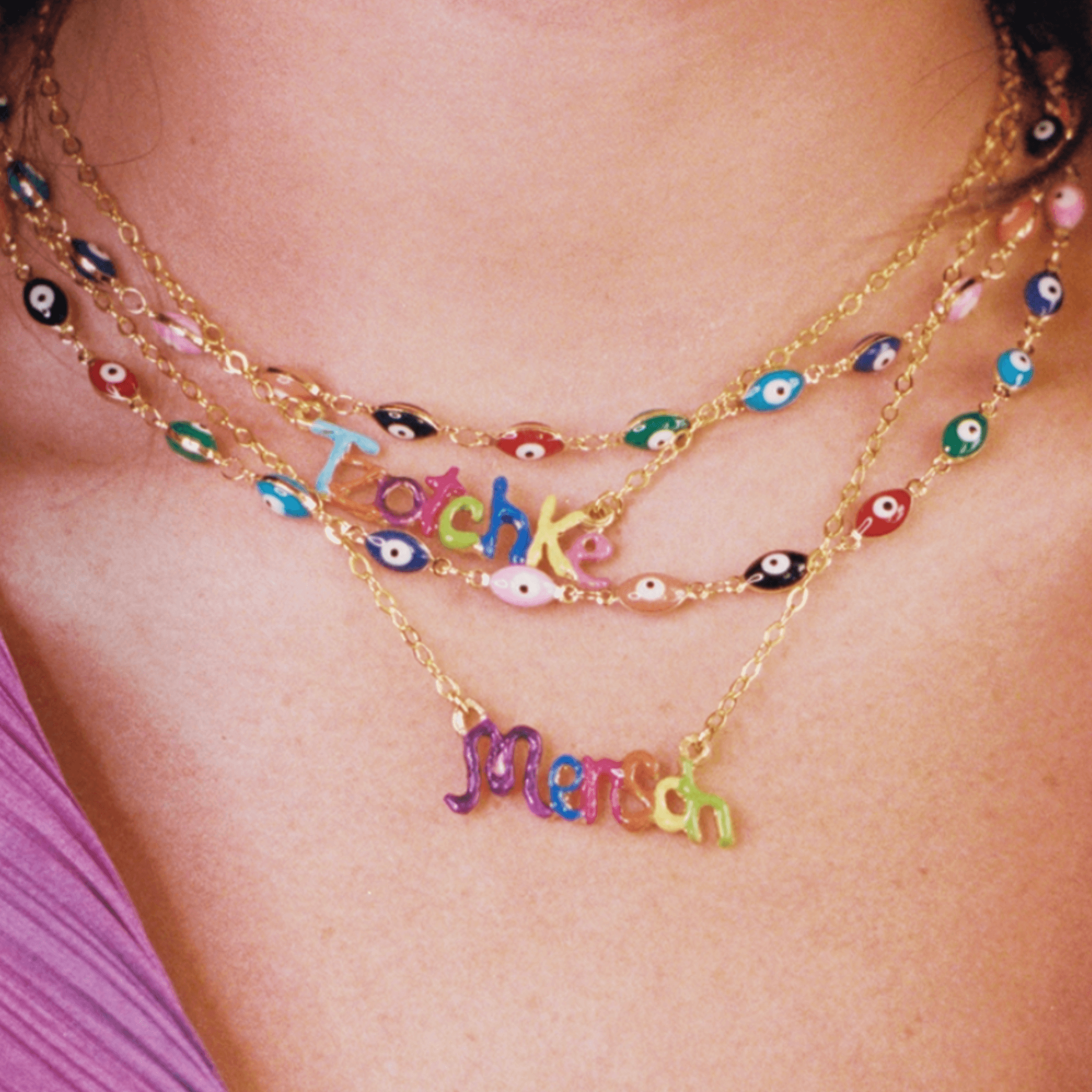 Layered gold necklaces featuring colorful enamel evil eye charms and playful words 'Tachke' and 'Merch'.