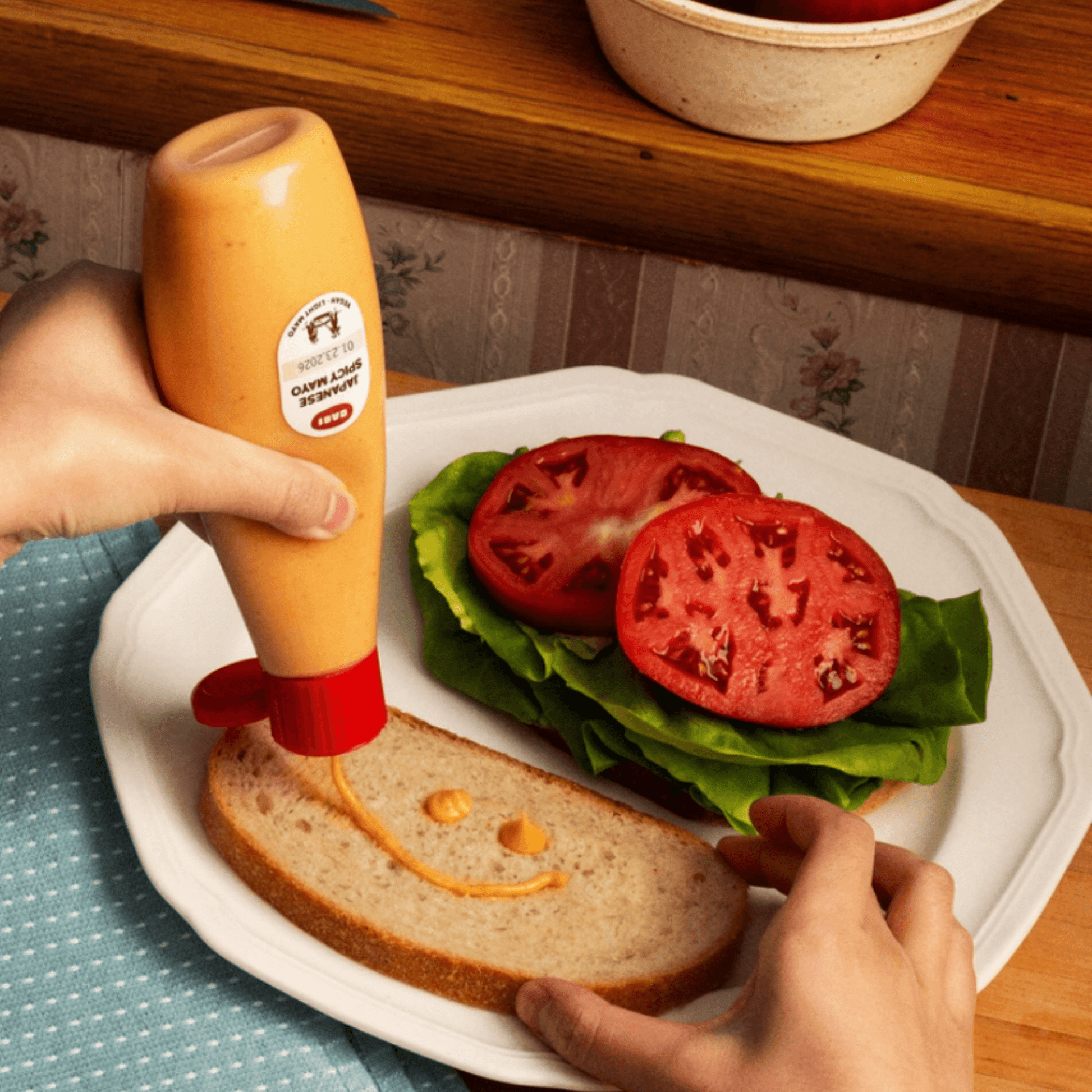 Hand squeezing Japanese spicy mayo onto a slice of bread with lettuce and tomato.