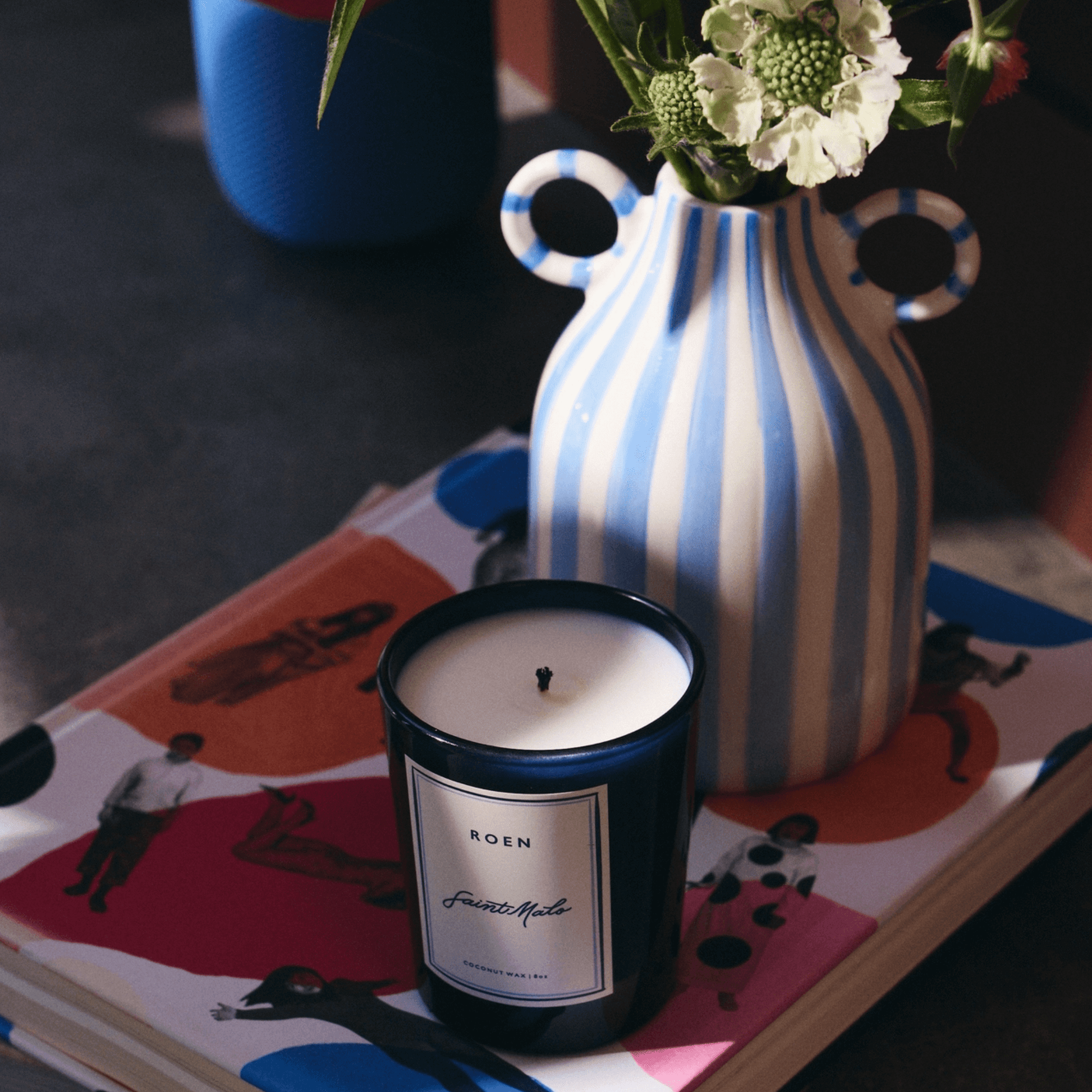 SAINT MALO candle by ROEN on a colorful magazine with a striped vase and fresh flowers.