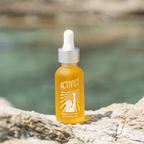 Precious Oils Active Infusion facial oil in dropper bottle on a rock by the sea, showcasing its vibrant color and natural setting.