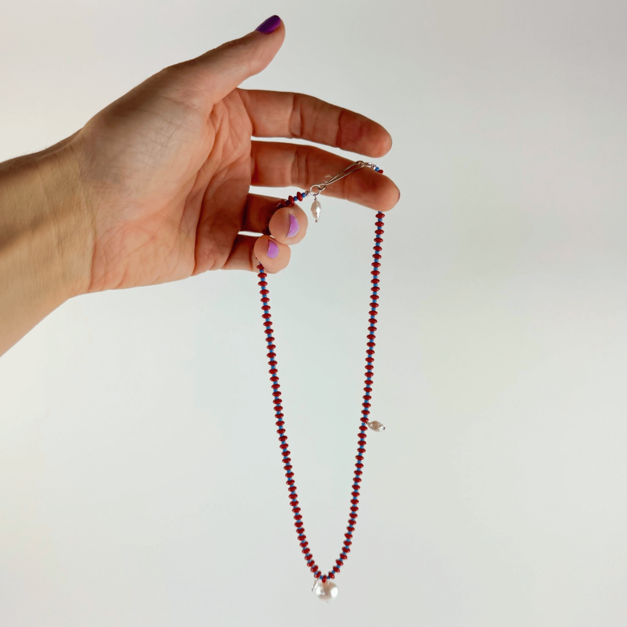 Hand holding Anni Necklace featuring red coral, blue seed beads, and pearls, showcasing its elegant design and craftsmanship.
