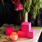 Colorful De Las Flores candle with pink packaging, surrounded by vibrant flowers on a marble table.