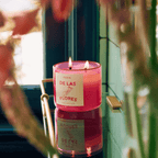 Pink DE LAS FLORES candle glowing on a reflective surface surrounded by florals, evoking Mexico City's flower market scent.