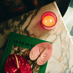Cozy scene featuring DE LAS FLORES candle on a marble table with a colorful book and vibrant pink leaves.