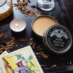 Sore Muscle Salve tin by Fat and the Moon among herbs and a candle, perfect for tension relief and soothing aches.