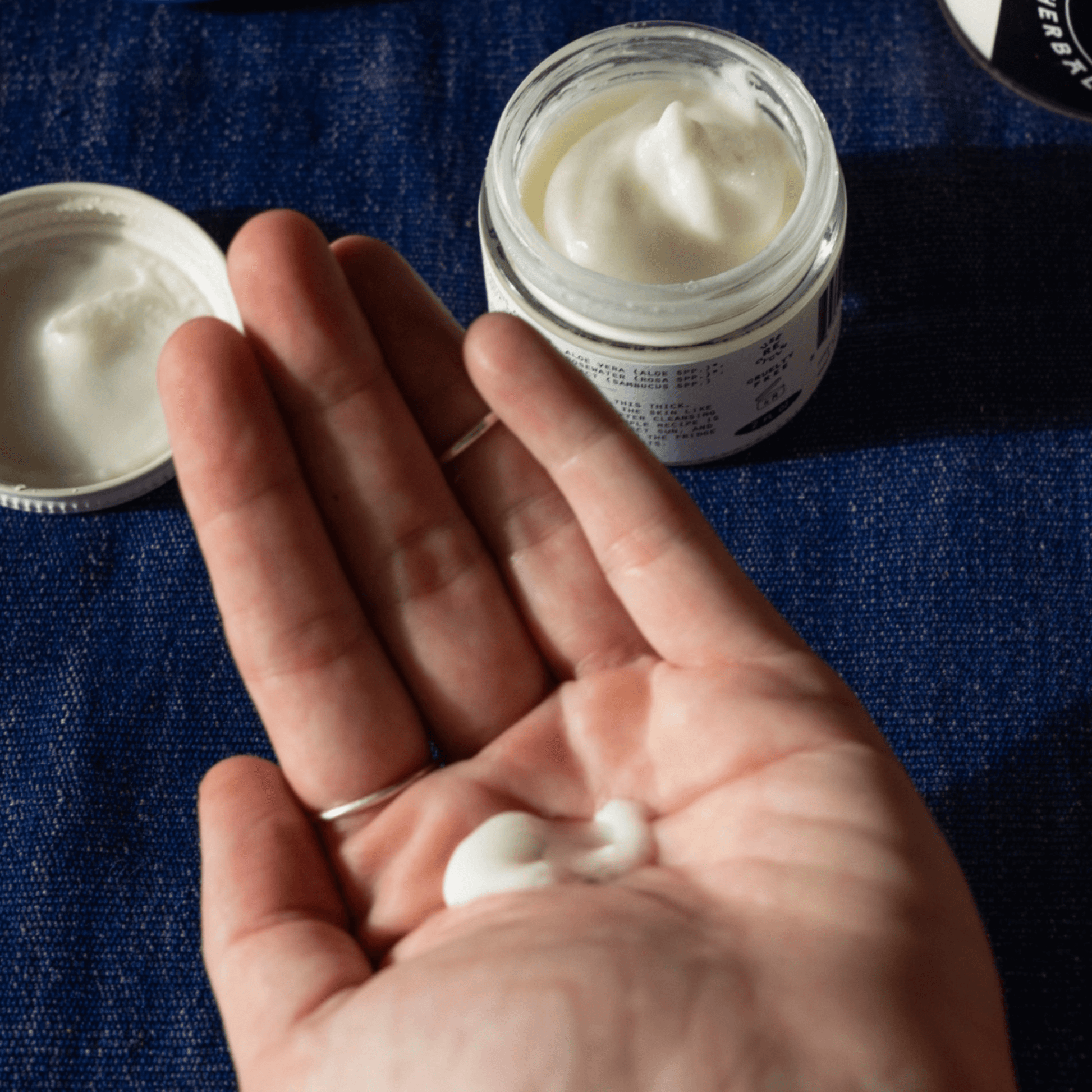 A hand holds a dollop of All Cream, showcasing its rich texture beside an open jar on a blue fabric background.