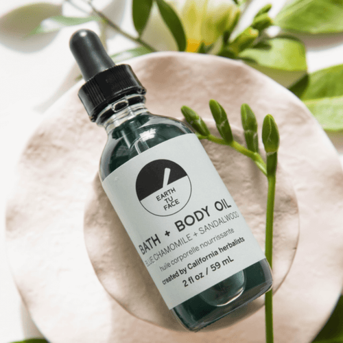 2 oz bottle of Earth Face Bath and Body Oil with blue chamomile and sandalwood, surrounded by natural elements.