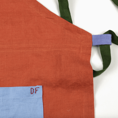 Close-up of orange linen apron featuring color-blocked corner and contrasting blue pocket and green straps.