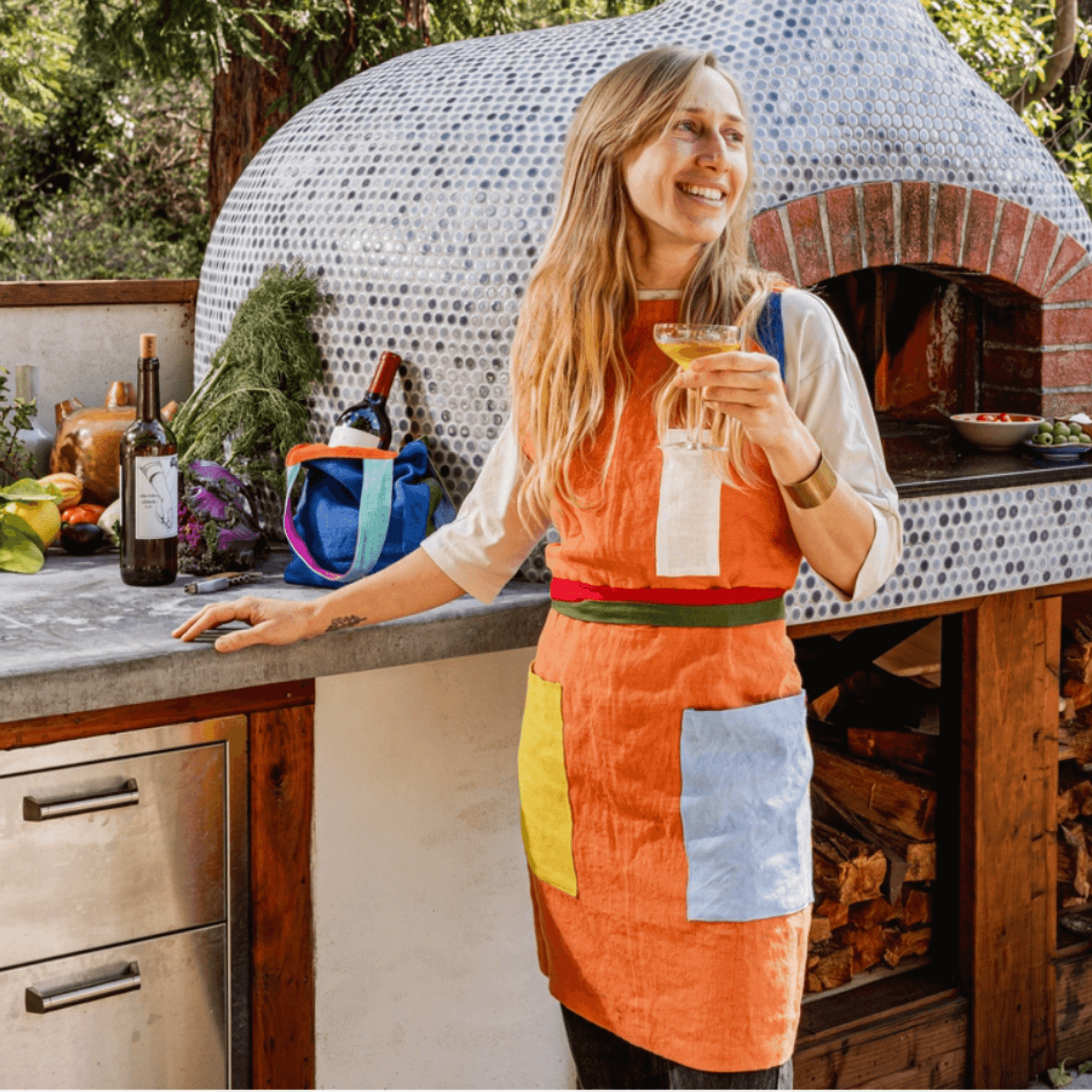 Woman wearing an orange linen apron with contrasting pockets, enjoying a drink in a rustic kitchen setting.