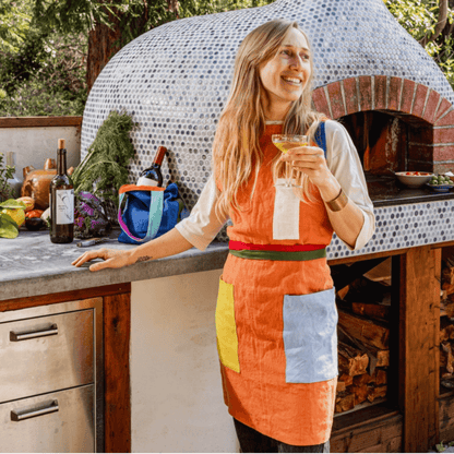 Woman wearing an orange linen apron with contrasting pockets, enjoying a drink in a rustic kitchen setting.