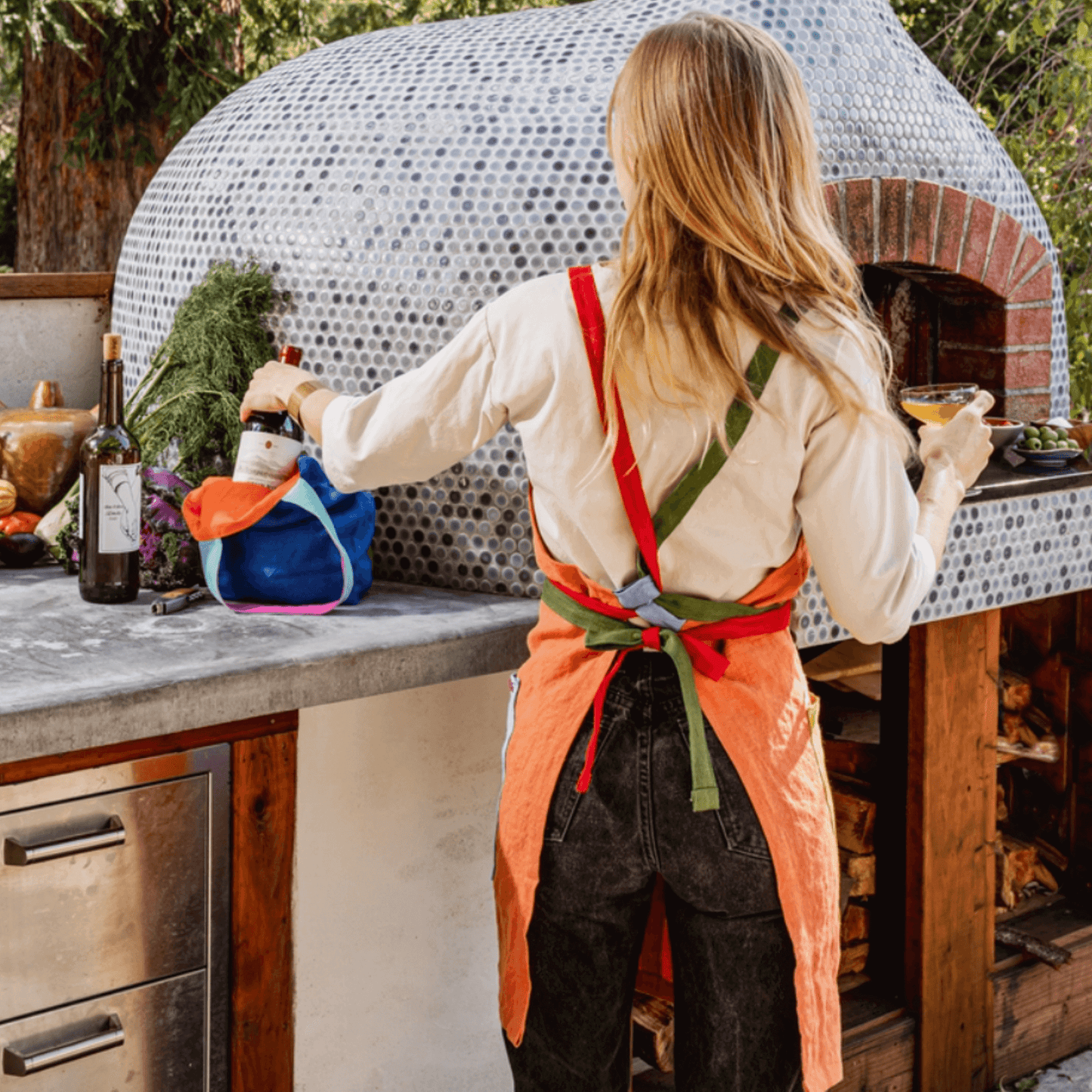 Person wearing an orange linen apron with colorful straps, preparing food by an outdoor oven.