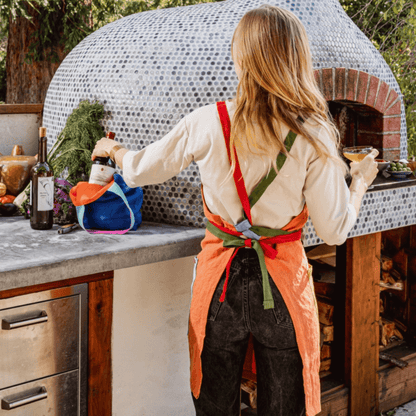 Person wearing an orange linen apron with colorful straps, preparing food by an outdoor oven.
