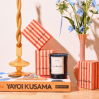 Elegant La Maison Rose candle on a vibrant table with striped boxes and flowers, exuding French countryside charm.