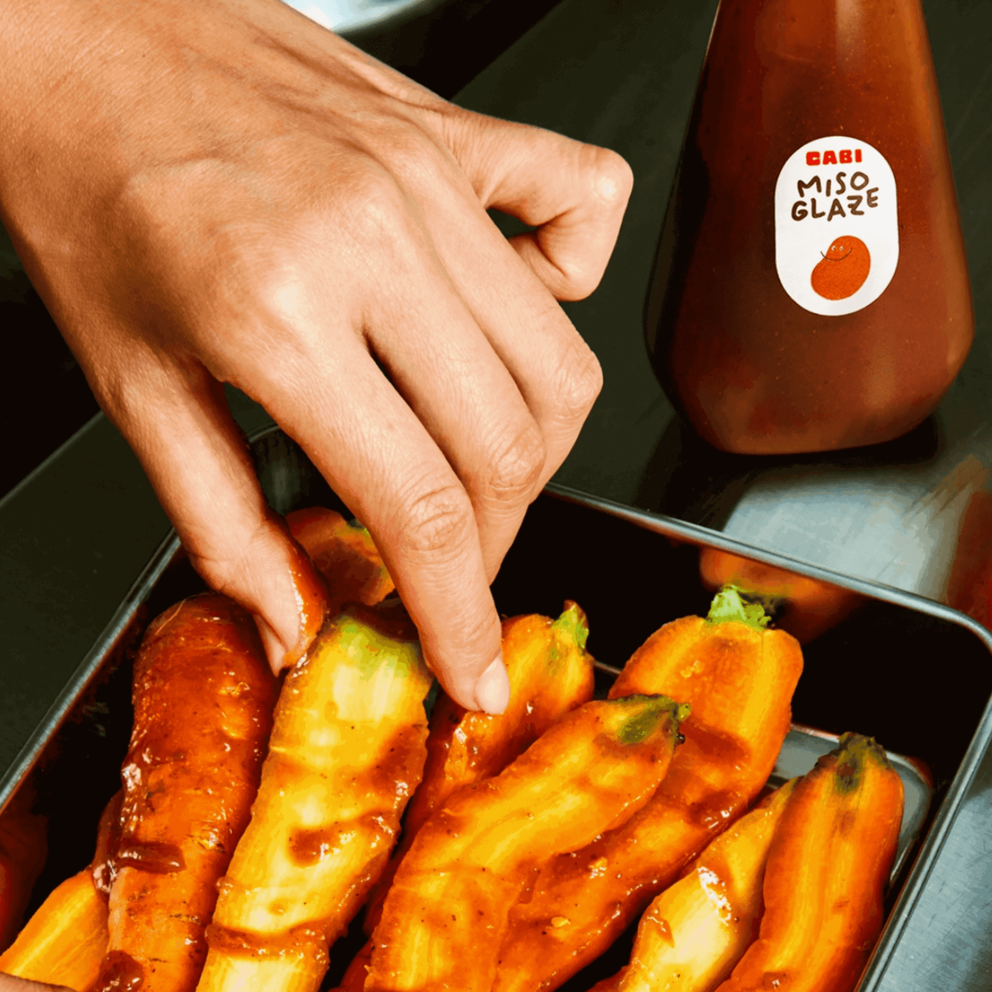 Hand drizzling Cabi Miso Glaze over grilled vegetables, showcasing bold umami flavors for home cooking.