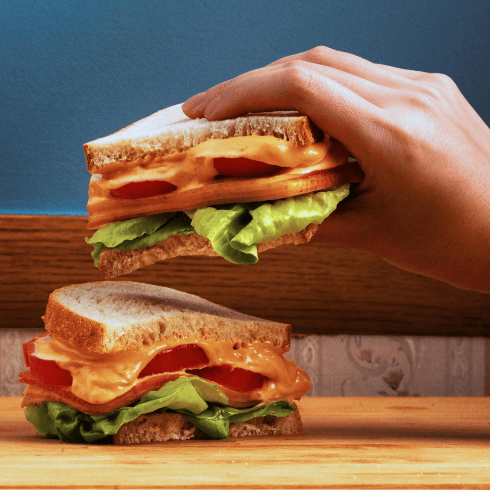 A hand holding a sandwich layered with spicy mayo, lettuce, and tomato on white bread, set against a colorful background.
